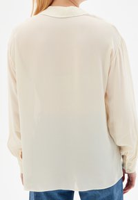 Femme portant un blouse longue à manches longues couleur crème, ample, assortie à un jean bleu, vue de dos contre un fond blanc.