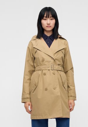 Femme aux cheveux noirs et lisses portant un trench-coat beige à double boutonnage avec une ceinture et un pantalon bleu sur un fond uni.