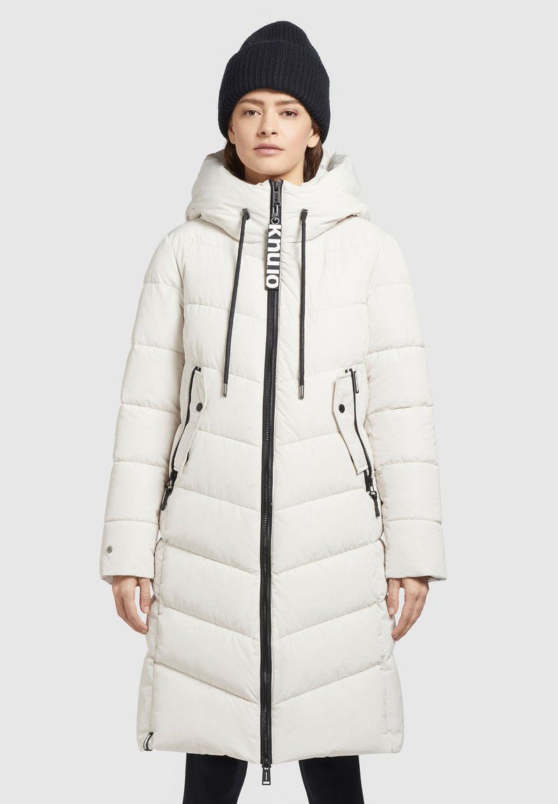 Witte pufferjas met een diagonaal gestikt patroon, voorrits, zijzakken en een zwart trekkoord label; gedragen met een zwarte beanie.