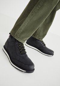 Botas negras con cordones, hechas de material texturizado, con cordones verdes y una suela de goma blanca. Llevadas con pantalones de pana verde olivo.