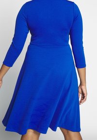 Une robe bleue à manches longues avec un corsage ajusté, une jupe évasée et un tissu à texture lisse, dotée d'un col rond et d'un ourlet à hauteur du genou.