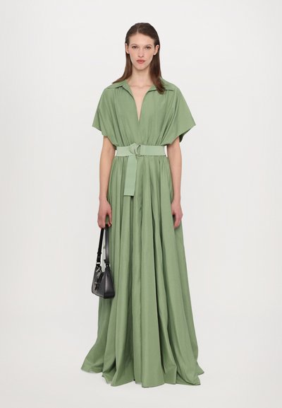 Vestido maxi verde com um decote em V profundo, mangas curtas em forma de capa, cintura ajustada com um cinto e uma saia fluida. Modelo segura uma bolsa preta.