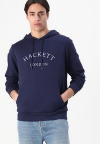 Námořnická modrá mikina s kapucí vyrobená z měkké tkaniny, s přední kapsou ve stylu "kangaroo", stahovací kapucí a stříbrně tištěným názvem značky "HACKETT LONDON."