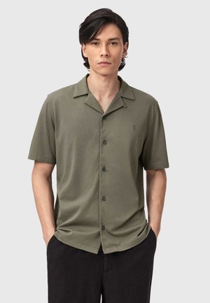 Junger Mann mit dunklem Haar, der ein olivgrünes kurzärmliges Hemd mit Knöpfen und schwarze Hosen trägt, Hände in den Taschen, neutraler Hintergrund.