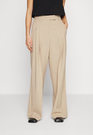 Femme portant un pantalon beige à jambes larges, avec des plis et des poches latérales, assorti à des chaussures noires et un haut noir sans manches.