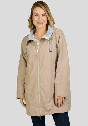 Femme souriante portant un manteau beige zippé avec une capuche grise sur un pantalon foncé, debout devant un fond gris clair uni.