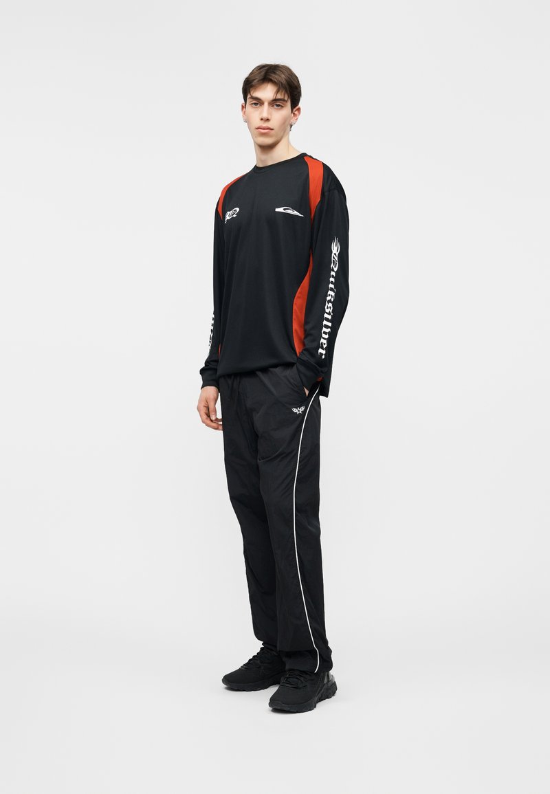 Άνδρας μοντέλο όρθιος, φορώντας μαύρο και κόκκινο μακρυμάνικο πουκάμισο με λευκά γράμματα στα μανίκια, μαύρο παντελόνι με λευκές ρίγες στα πλαϊνά και μαύρα αθλητικά παπούτσια.