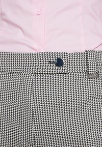 Pantaloni a mosaico houndstooth bianco e nero con frontale piatto, caratterizzati da un bottone blu e un'etichetta metallica Hugo sulla cintura. Camicia rosa a maniche lunghe sopra.