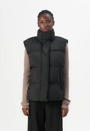 Black puffer vest with high collar, quilted design, and a logo patch. Features two front pockets and wide armholes. Textured inner layer visible.