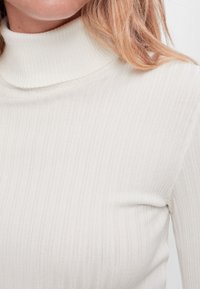 Zbliżenie na osobę ubraną w białą, prążkowaną sweter-karkot z widocznymi jasnobrązowymi włosami po prawej stronie.