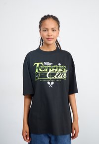 Liela melna t-krekls, kas izgatavots no kokvilnas, ar neona zaļu "Nike Tennis Club" grafiku, kurā iekļauti tenisa motīvi un dibināšanas datums.