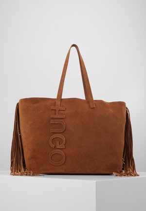 Braune Wildleder-Shopper-Tasche mit doppelten Griffen, Fransen-Details an den Seiten und vertikal geprägtem "QUH" auf der Vorderseite, auf weißer Oberfläche präsentiert.