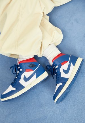 Niebiesko-biało-czerwone wysokie buty sportowe Nike noszone z białymi skarpetkami i beżowymi spodniami na niebieskim dywanowym podłożu.