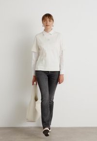 Polo shirt bianco a maniche corte sopra una maglietta a maniche lunghe, abbinato a jeans grigio scuro e scarpe nere senza lacci, mentre tiene una borsa tote bianca.