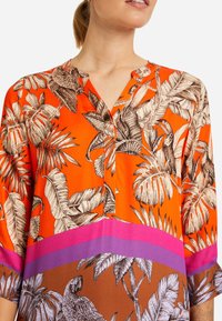 Blusa colorata in tessuto arancione con motivi di foglie tropicali neri e bianchi. Presenta un collo a V, maniche a tre quarti e dettagli in rosa.