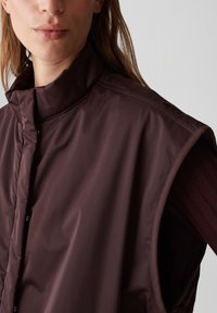 Bruine, mouwloze vest gemaakt van glanzend, lichtgewicht materiaal; heeft een hoge kraag, drukknoopjes en een gladde textuur met minimale details.