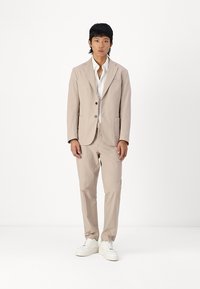 Beige suit with a textured finish, consisting of a jacket with two buttons and matching pants. White shirt underneath, paired with white sneakers.