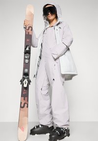 Costum de schi violet cu glugă, având fermoare și buzunare, purtat cu bocanci de schi negri; ținând schiuri maro cu legături negre.