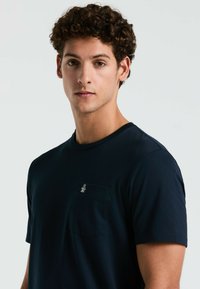 Marineblaue Baumwoll-T-Shirt mit Rundhalsausschnitt, kurzen Ärmeln und einer linken Brusttasche mit einem kleinen Pinguin-Logo. Glatte Textur.