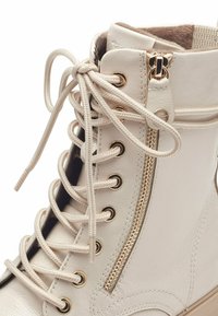 White faux leather ankle boot with flat laces, gold eyelets, side zipper, and textured finish. Cushioned collar and padded interior.