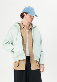 Mint green zip-up hoodie with a front pocket, layered over a tan button-up shirt. The model wears a light blue cap with a logo.