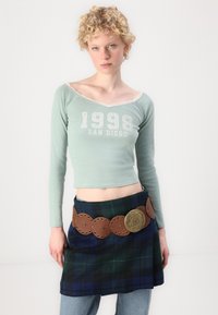 Minto žalia ilgomis rankovėmis palaidinė su nėriniuota apdaila ir spausdintu užrašu "1998 San Diego", derinama su mėlyna ir žalia kare dešra sijonu, kuriame yra rudas papuoštas diržas.
