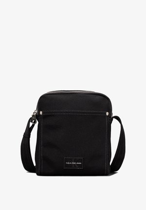 Sac bandoulière noir Calvin Klein Jeans avec poche avant, coutures blanches, fermeture éclair et sangle réglable.