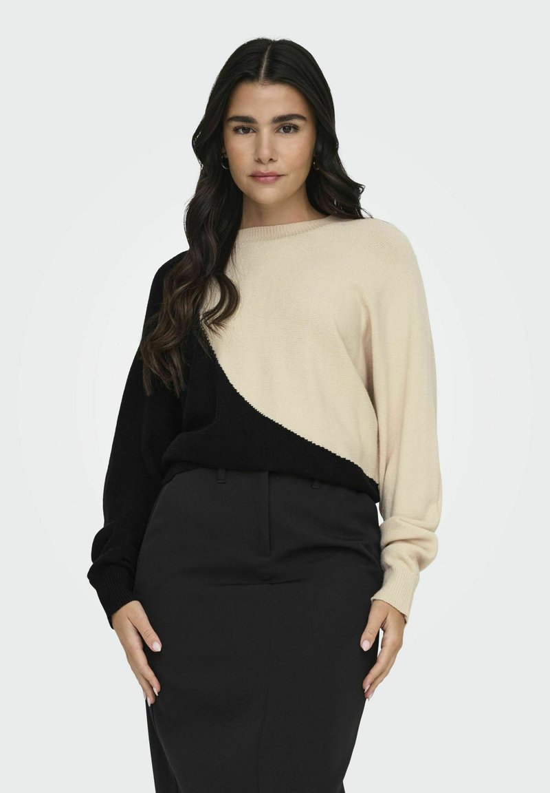 Maglione lavorato a maglia con colori crema e nero, caratterizzato da un design a spacco diagonale. Maniche lunghe e polsini a coste, abbinato a pantaloni scuri.