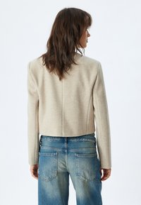 Chaqueta recortada de mezcla de lana beige con hombros estructurados y costuras rectas, combinada con pantalones de mezclilla azul claro desgastados.