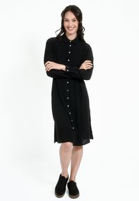 Must üleval nööbitud must kleit, millel on krae, pikad varrukad ja seotud talje. Sile kangas, põlvekõrgune disain, koos mustade sametist tossudega.