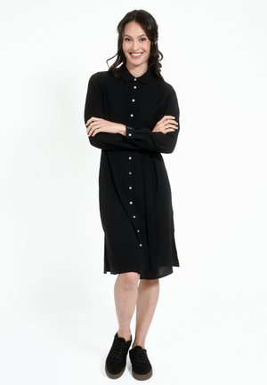 Schwarzes Hemdkleid mit Kragen, langen Ärmeln und gebundenem Taillenbündchen. Glatte Stoff, knielanges Design, kombiniert mit schwarzen Wildleder-Sneakern.