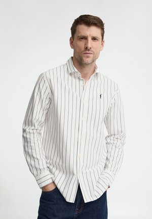 Uomo che indossa una camicia bianca a bottoni con sottili righe verticali nere e jeans blu scuro, in piedi con le mani in tasca davanti a uno sfondo bianco.