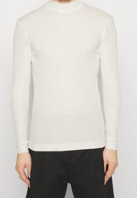 Suéter de cuello alto de manga larga en color beige claro. Textura de punto, diseño ajustado, puños y dobladillo acanalados. Combinado con pantalones de traje negros.
