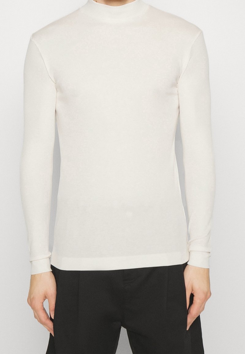 Suéter de cuello alto de manga larga en color beige claro. Textura de punto, diseño ajustado, puños y dobladillo acanalados. Combinado con pantalones de traje negros.