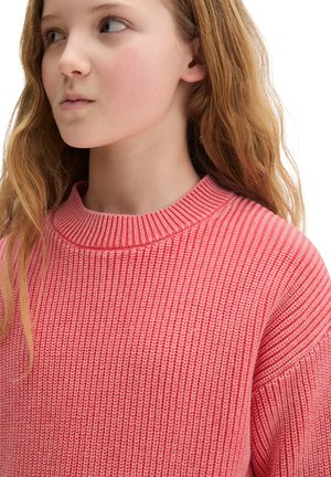 Jeune fille aux longs cheveux châtain clair portant un pull en tricot rose texturé regardant sur le côté sur fond blanc.