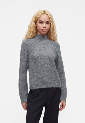 Jeune femme aux longs cheveux blonds bouclés, portant un pull gris à col roulé en maille et un pantalon noir, debout devant un fond uni.