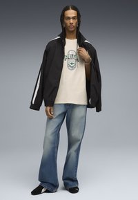 Sort jakke med hvide striber, lys beige t-shirt med grafisk print, blå flagrende jeans og sorte sko.