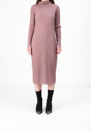 Vrouw die een lange mouw, geribbelde, mauve midi-jurk draagt met zwarte soklaarzen met puntige neuzen, staand op een witte achtergrond.