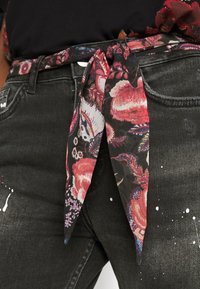 Pás z látky s květinovým vzorem, na černém pozadí růžové a fialové květiny. Uvazuje se do uzlu, nošený přes tmavé džíny poseté barvou.