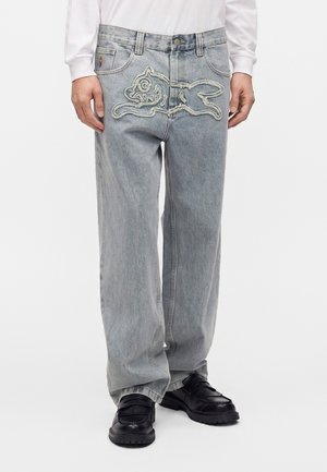 Homme portant un jean large gris clair avec un dessin cousu d’un animal de dessin animé à l’avant et des chaussures noires, debout devant un fond blanc.