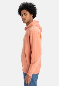 Sudadera con capucha en color melocotón, fabricada en un tejido suave, con un bolsillo tipo canguro, cierre con cordón y puños acanalados. Diseño sencillo.