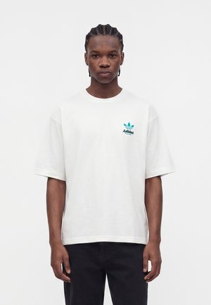 Ung mand med flettet hår står med fronten mod kameraet iført en hvid Adidas Originals t-shirt med turkis logo og sorte bukser.