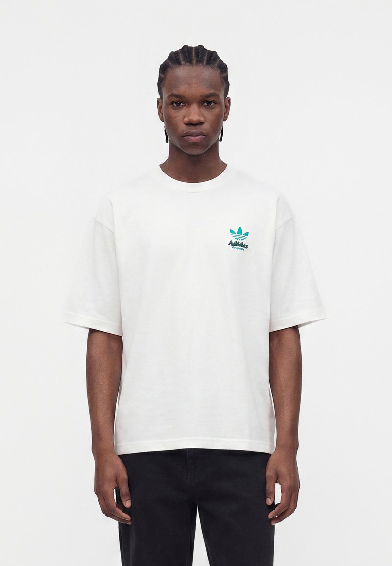 Junger Mann mit geflochtenem Haar steht frontal und trägt ein weißes Adidas Originals T-Shirt mit türkisem Logo und schwarze Hose.