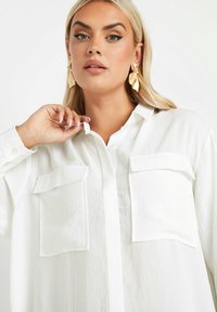 Camicia bianca con bottone, dotata di due tasche sul petto, realizzata in un tessuto leggero. Presenta un colletto e una vestibilità comoda. Capelli lasciati sciolti.