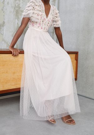 Kvinde iført en hvid, broderet, gennemsigtig blomsterkjole med pufærmer, siddende på et træskab mod en betonvæg.