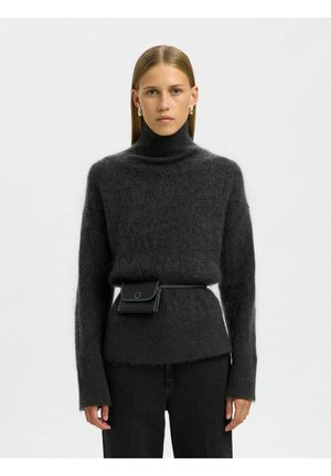 Donna con capelli biondi lisci che indossa un maglione dolcevita peloso grigio scuro, pantaloni neri, orecchini a cerchio argentati e una borsa a cintura nera.