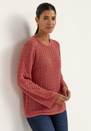 Femme aux cheveux foncés portant des boucles d'oreilles dorées et un pull rose texturé à manches longues, debout contre un mur blanc, les mains jointes.