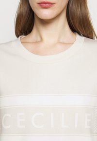 Jasno-beżowy sweter z okrągłym dekoltem, miękką teksturą i poziomym wzorem w paski z nazwą "CECILIE" napisanymi grubymi białymi literami.