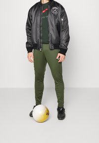 Osoba v černé bundě Paris Saint-Germain, zelených kalhotách a černých kopačkách Nike stojí se fotbalem u nohou.