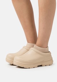 Zoccoli beige in gomma liscia con un risvolto a maglia testurizzata, punta arrotondata e suola robusta per una buona aderenza.
